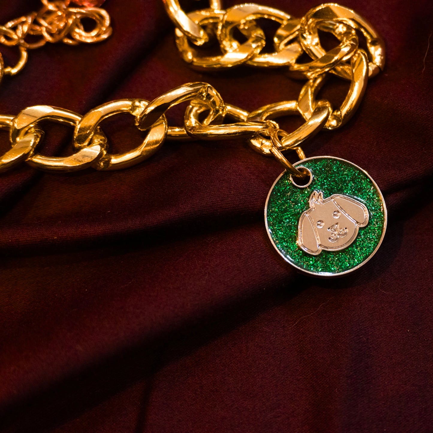 Gold pet necklace with green glitter dog-face charm displayed on a maroon cloth background