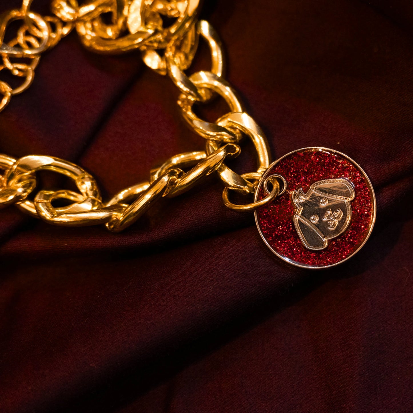 Gold pet necklace with red glitter dog-face charm displayed on a maroon cloth background
